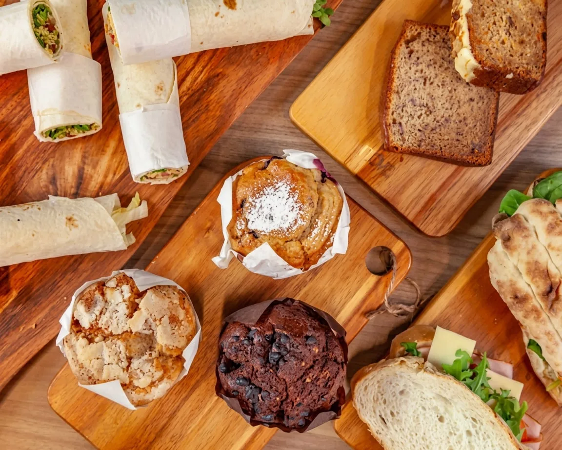 Wraps, muffins, banana bread and filled rolls laid out across wooden boards, ready to box for catering